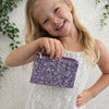 Young girl holding a purple floral pouch against a white textured background with green leaves.