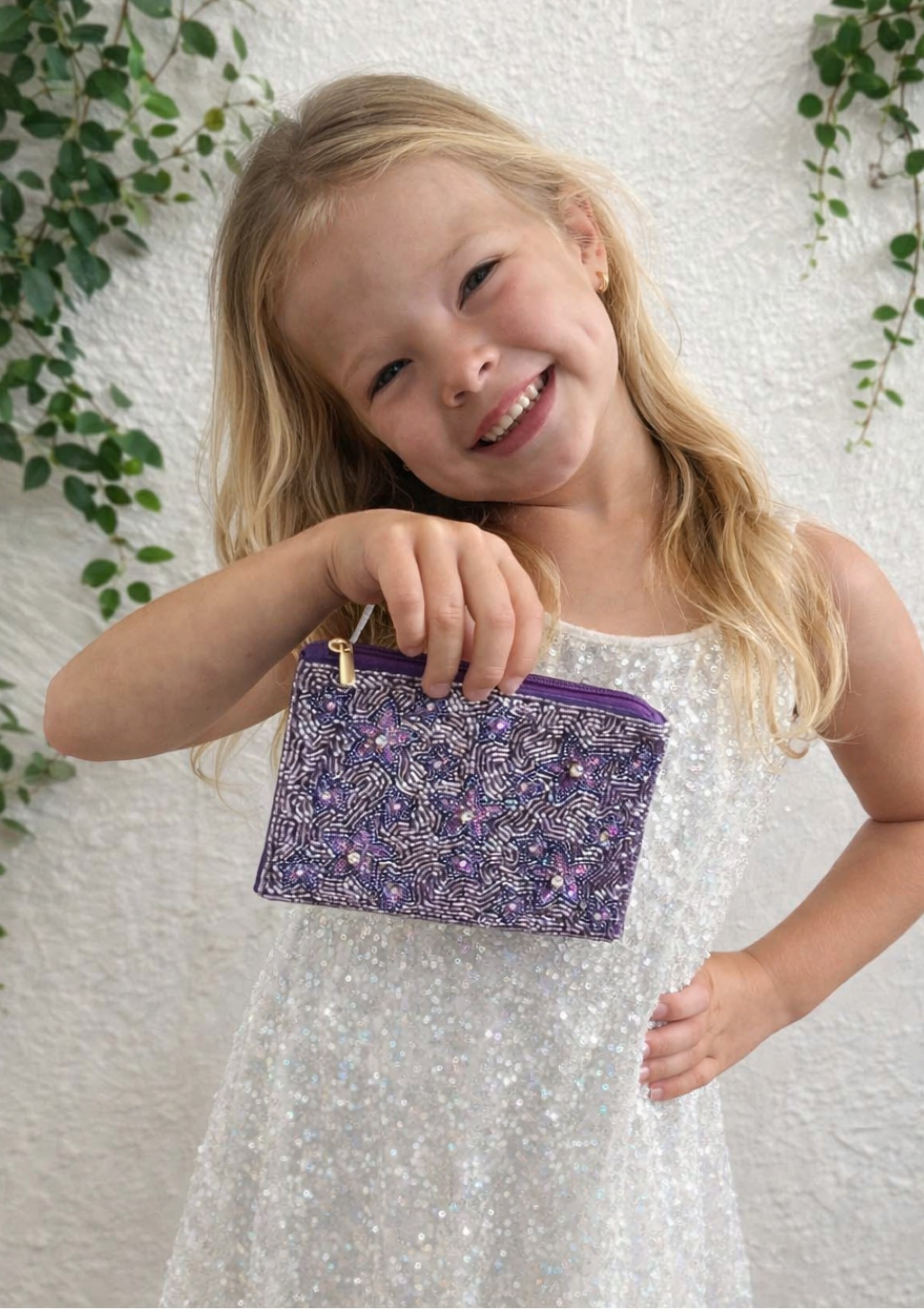 Young girl holding a purple floral pouch against a white textured background with green leaves.